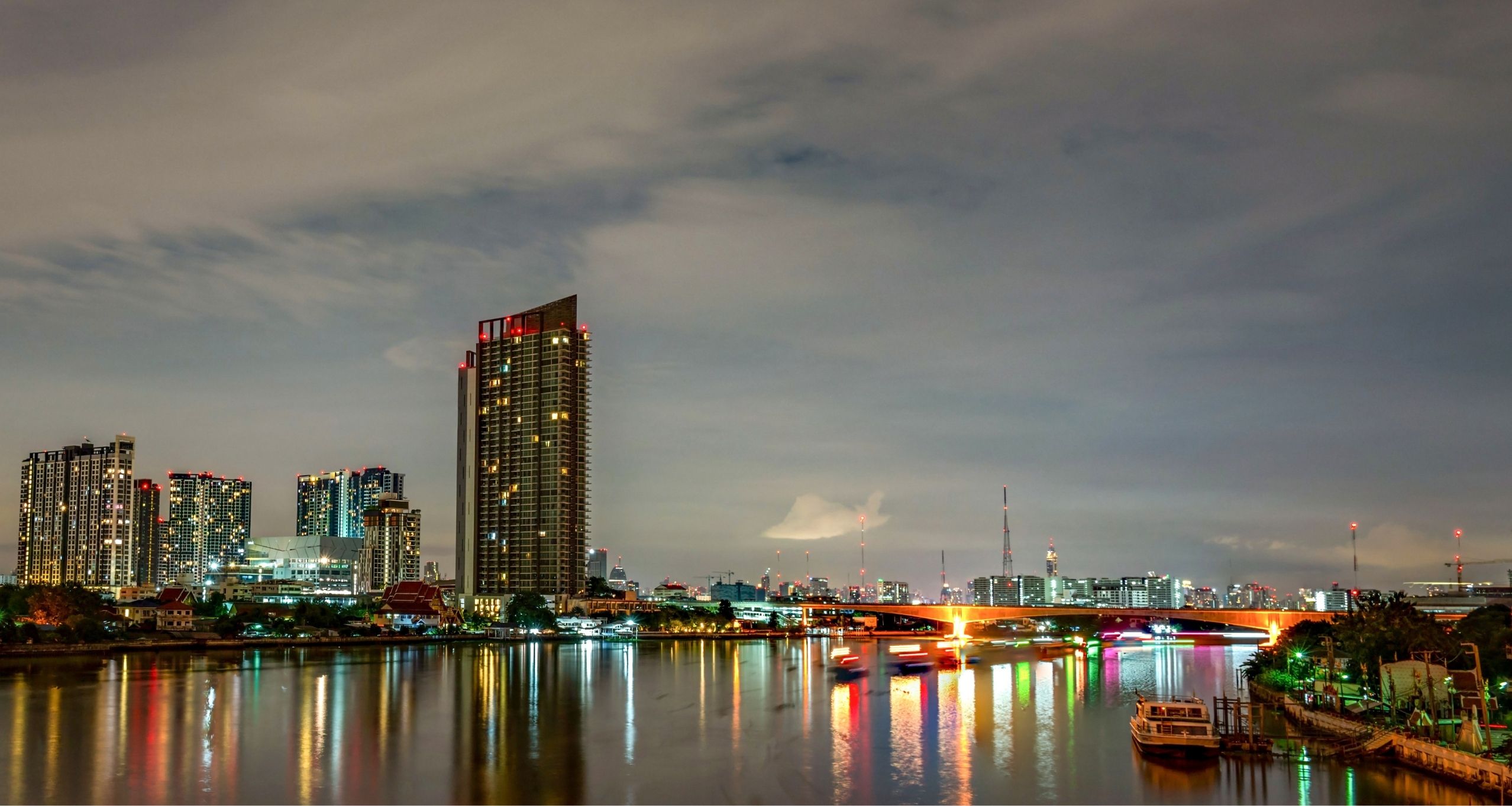 Night city skyline along a calm river, tall illuminated buildings and a bright bridge reflecting in the water at dusk.